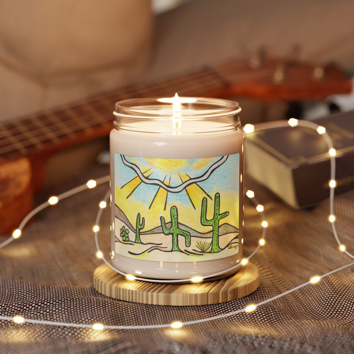 A lit candle in a clear glass jar sits on a round wooden coaster, surrounded by warm white fairy lights. The jar features a colorful desert landscape illustration with green cacti, a yellow sun, and a blue sky. A beautiful representation of the Southwest desert.