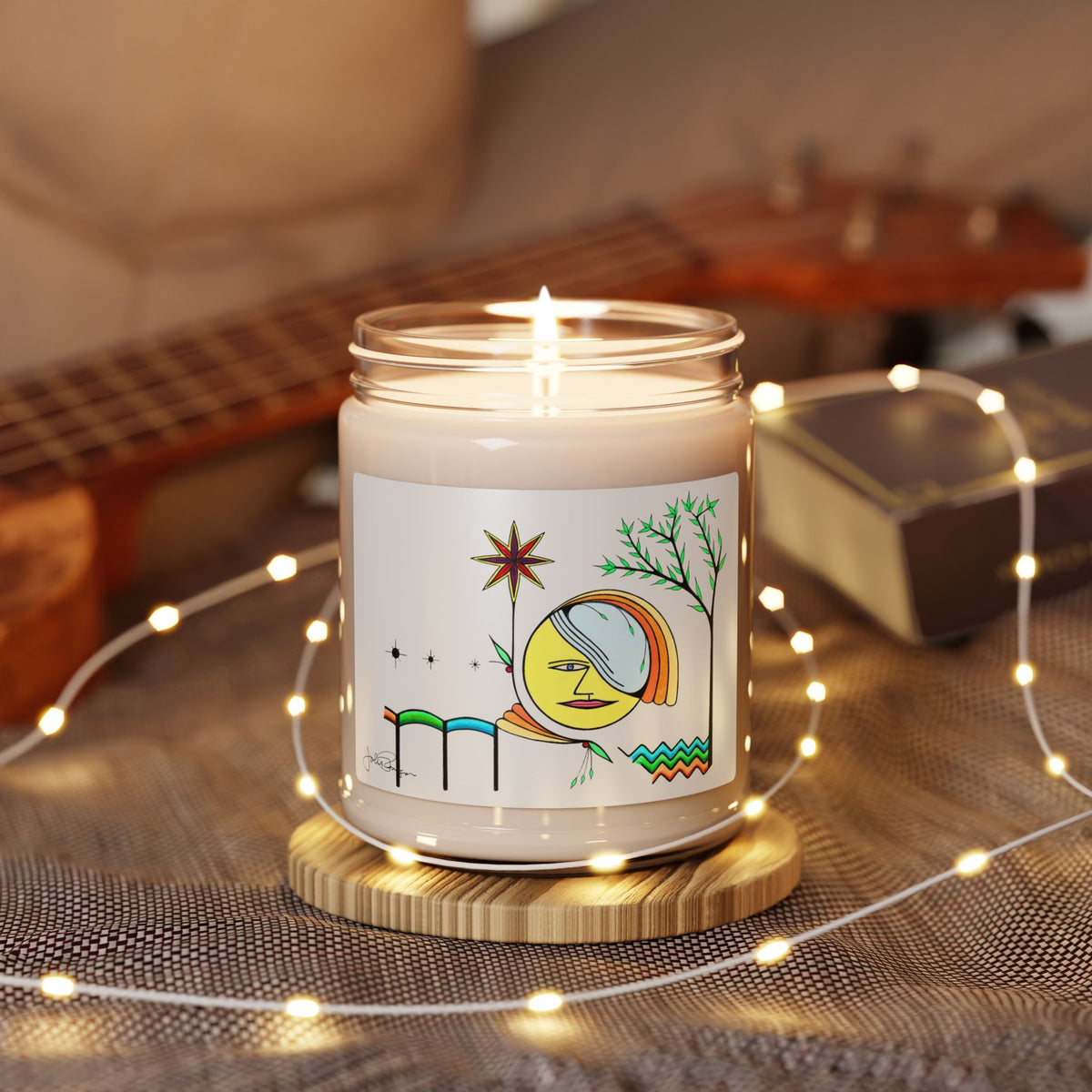 A lit candle in a clear glass jar sits on a round wooden coaster, surrounded by warm fairy lights. The candle has a white label with a whimsical illustration of a crescent moon, stars, and green branches.
